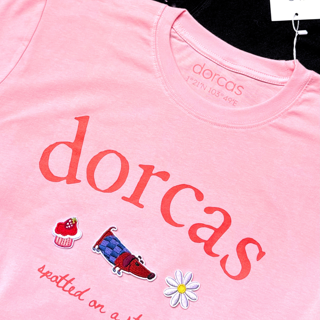 Pink t-shirt with 'dorcas' branding and graphics, displayed with a black t-shirt.