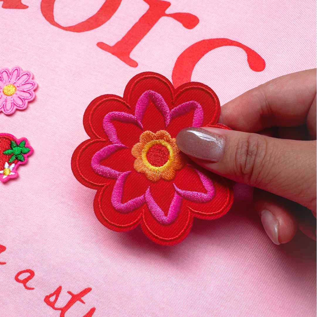 Hand holding a red flower-shaped patch over a pink fabric background with visible text.